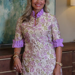 A smiling blonde woman wearing a short-sleeve floral dress with purple ruffled trim and a purple high neck, standing indoors in front of a textured wall and wooden cabinet by Kim Lolley Designs