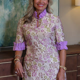 A smiling blonde woman wearing a short-sleeve floral dress with purple ruffled trim and a purple high neck, standing indoors in front of a textured wall and wooden cabinet by Kim Lolley Designs